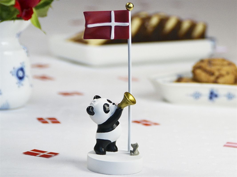 Panda med trompet og dansk flag på bordflag, dekoreret med kager og vase i baggrunden.